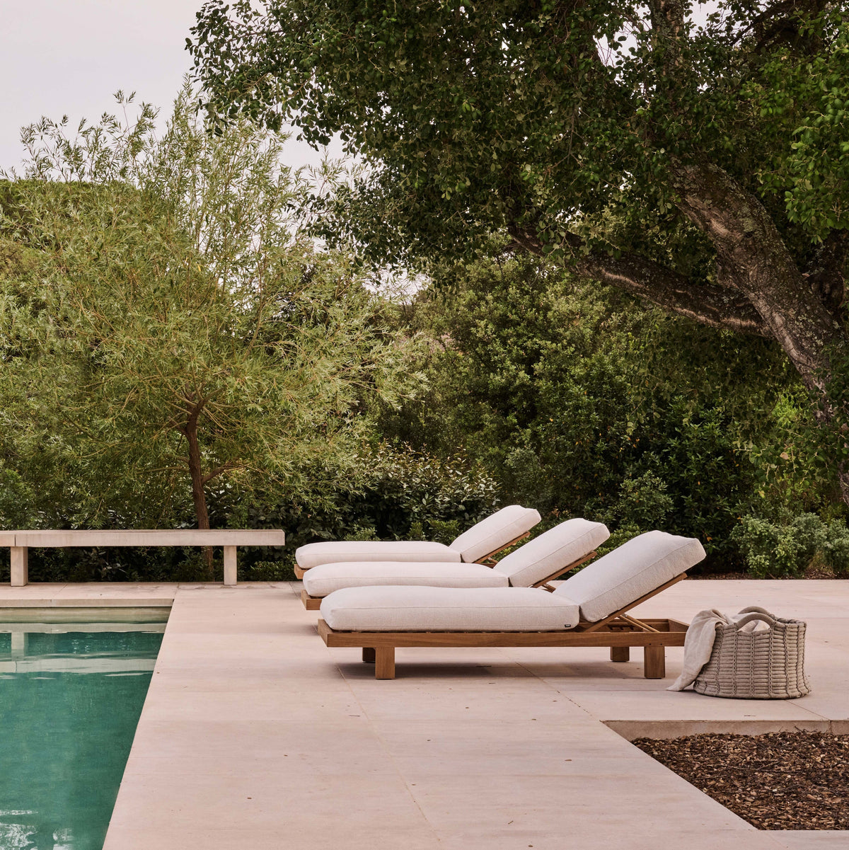 Tribù Pure Sonnenliegen aus Teak mit hellen Auflagen an einem Pool unter Bäumen