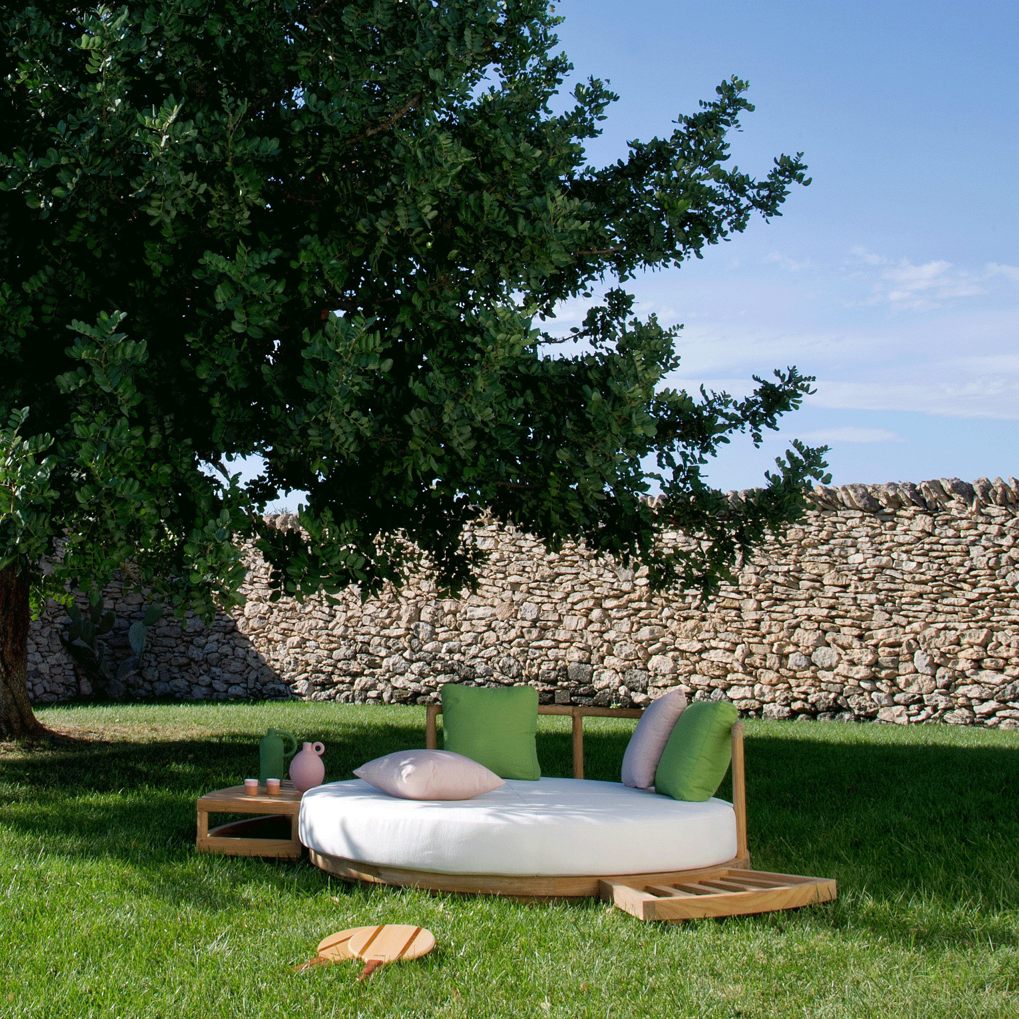 Unopiu Lounge Insel Pevero aus rundem Teakholz. Liegepolster in Acrylstoff weiß. Das Daybed steht im Rasen unter einem Haselnussbaum.