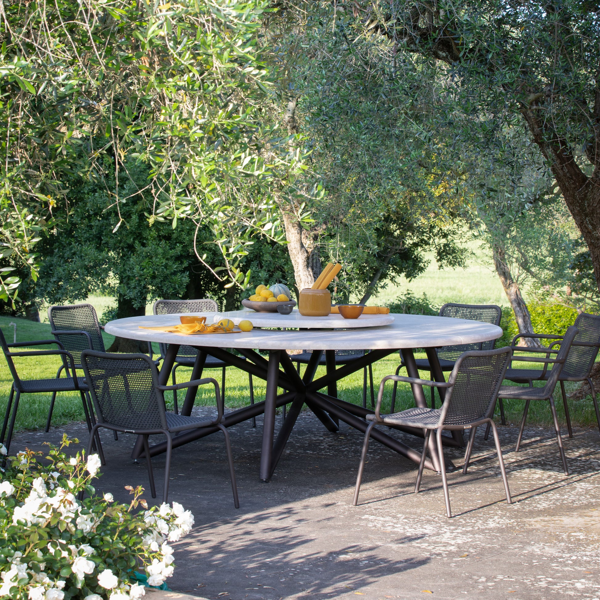 Der HERMU Gartentisch in einem eleganten Garten. Der Tisch besteht aus hochwertigem Peperino-Naturstein und hat eine drehbare Platte in der Mitte.