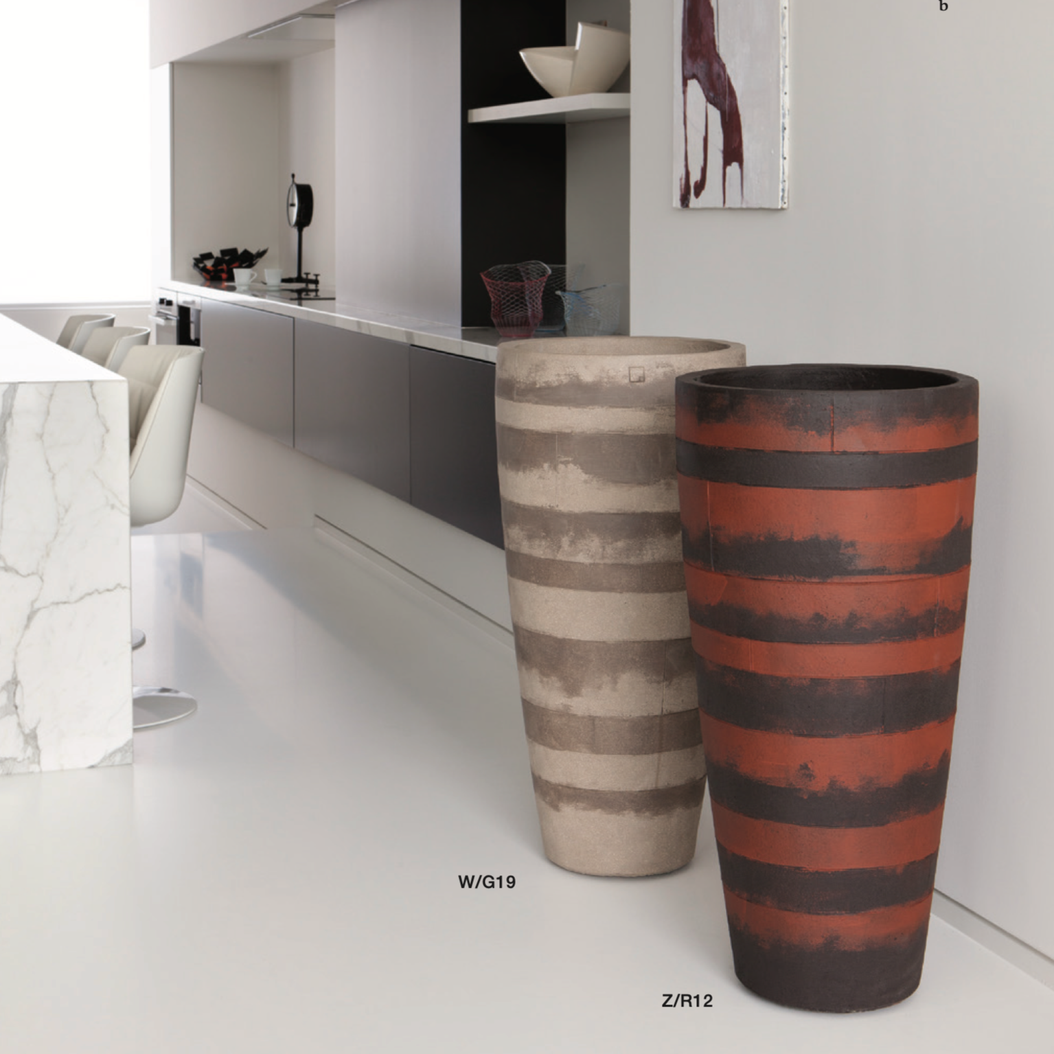 Zwei Atelier Vierkant Blumentröge eines in schwarz mit roten wagerechten Streifen, das 2 Gefäß ist weiss mit grauen wagerechten Streifen. Beide Gefäße stehen im Wohnbereich nebeneinander vor einer weißen Wand.