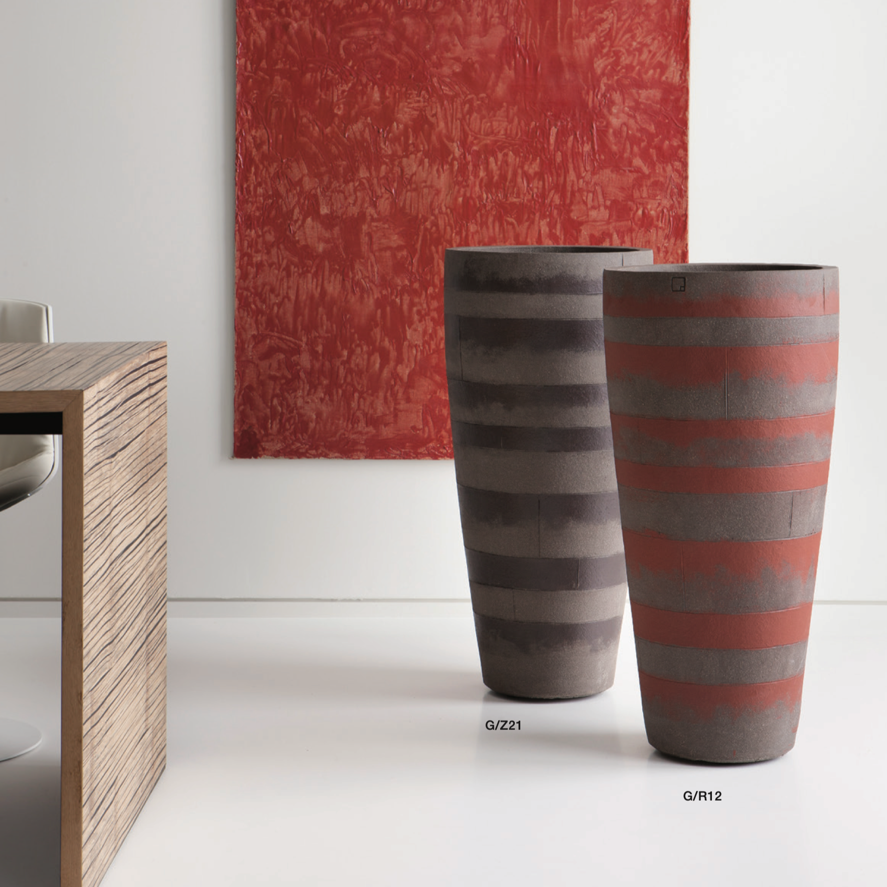 Zwei Atelier Vierkant Blumentröge eines in schwarz mit roten wagerechten Streifen, das 2 Gefäß ist weiss mit grauen wagerechten Streifen. Beide Gefäße stehen im Wohnbereich nebeneinander vor einer weißen Wand. 