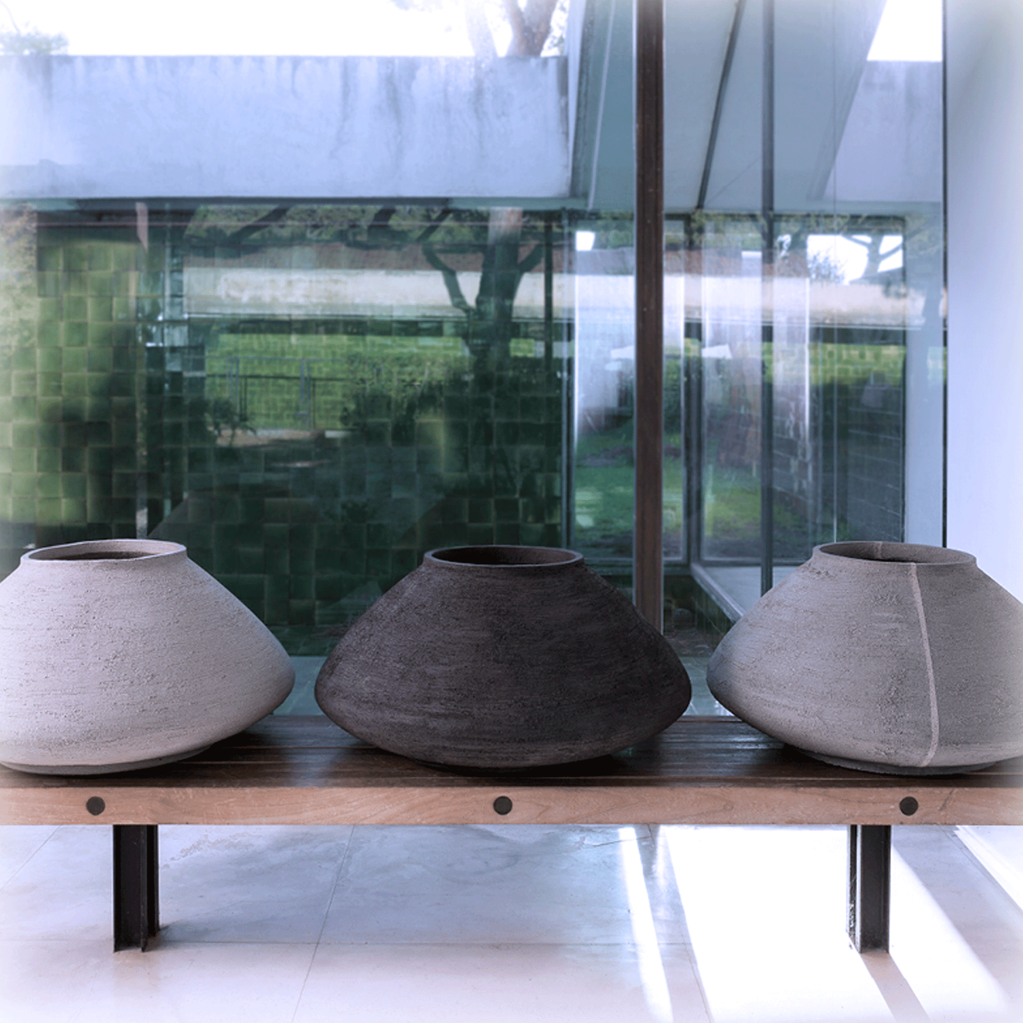 Drei Atelier Vierkant Vasen LRC70 stehen auf einer Bank mit einer Teakholz Sitzfläche. Von links nach rechts steht ein weißes, schwarzes und graues LRC70 Gefäß. 