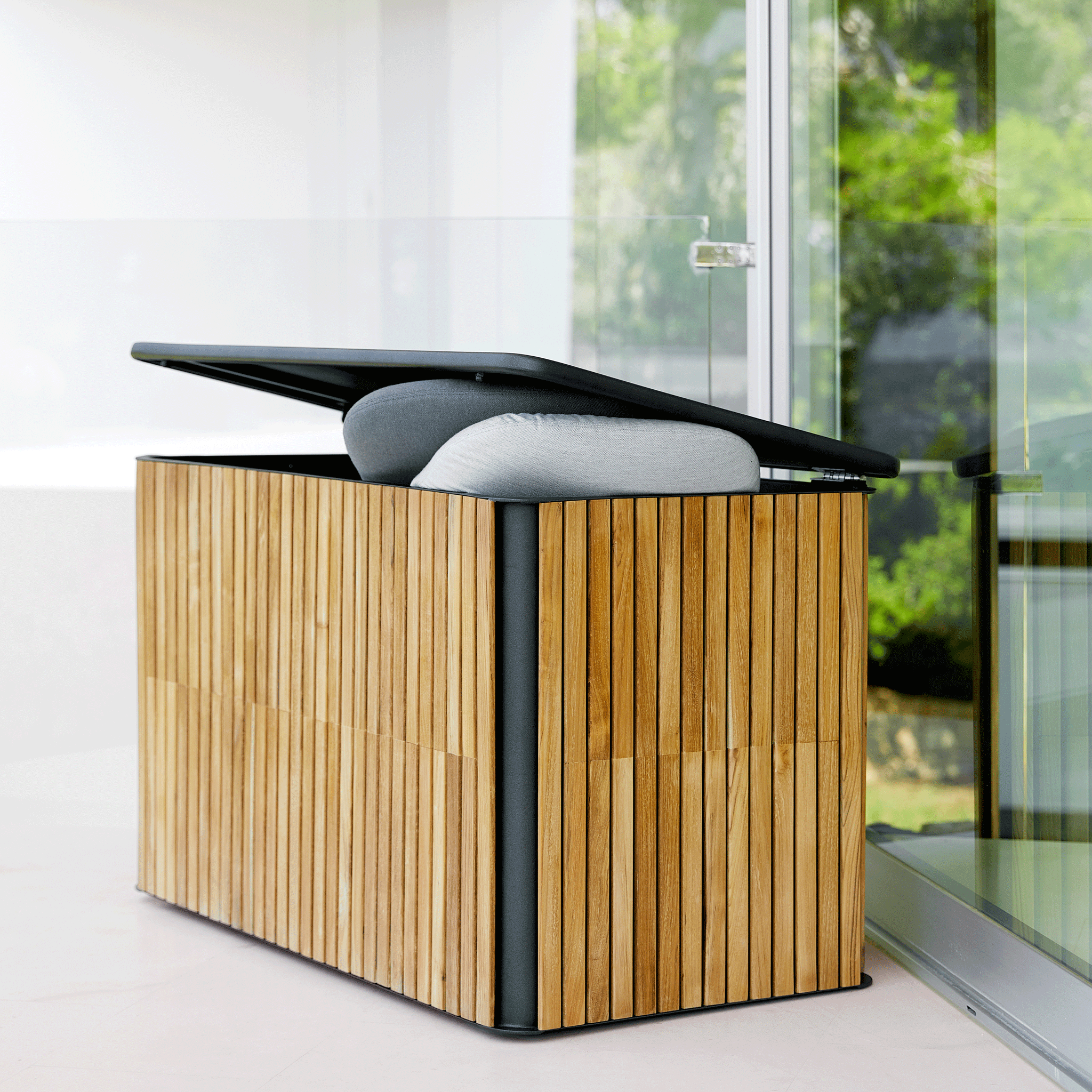 Die Combine Kissentruhe besteht aus einem Aluminiumgestell mit einer Teakholz Verkleidung. Die Kissenbox steht an einem sonnigem Tag auf einer modernen hellen Terrasse vor einer großen Fensterwand.Der Deckel ist leicht geöffnet und zwei Kissen in hell grau und grau erscheinen hervor.