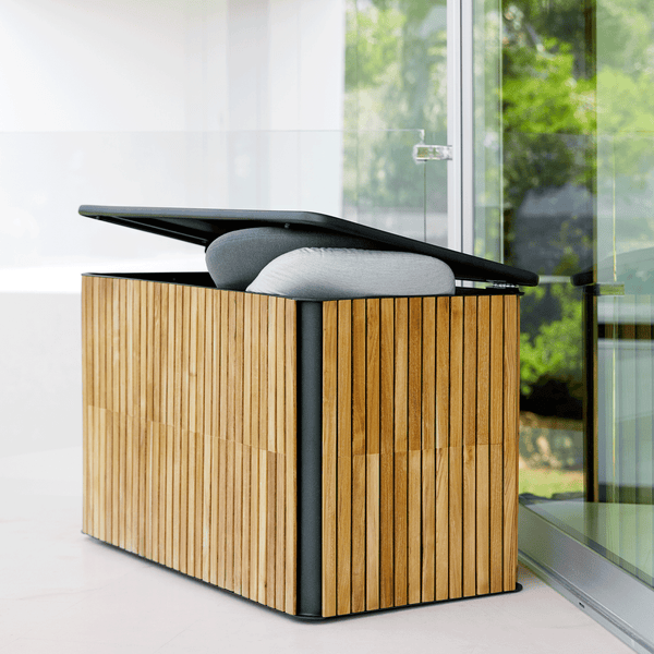 Die Combine Kissentruhe besteht aus einem Aluminiumgestell mit einer Teakholz Verkleidung. Die Kissenbox steht an einem sonnigem Tag auf einer modernen hellen Terrasse vor einer großen Fensterwand.Der Deckel ist leicht geöffnet und zwei Kissen in hell grau und grau erscheinen hervor.