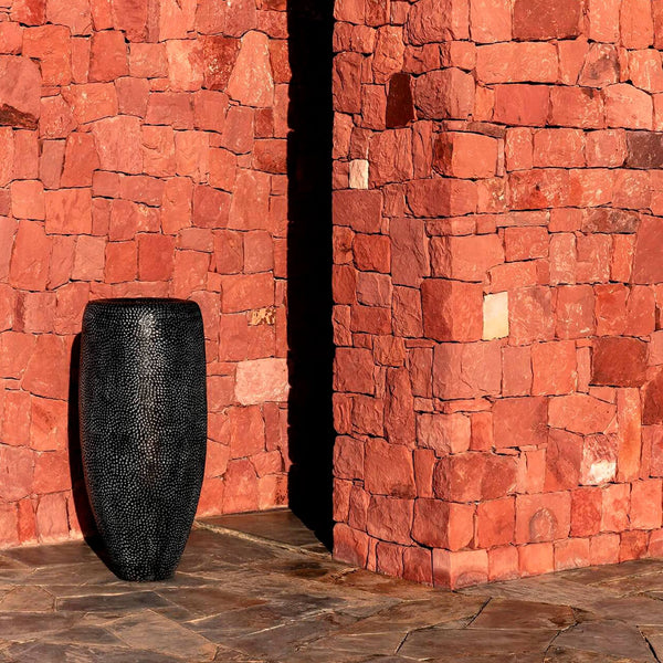 Vor einer roten Ziegelwand steht die Domani Vase BATUR aus Terracotta. Das Pflanzengefäß ist ein Designobjekt in schwarzer metallic Optik. Diese Blumenvase misst eine Höhe von 120cm.