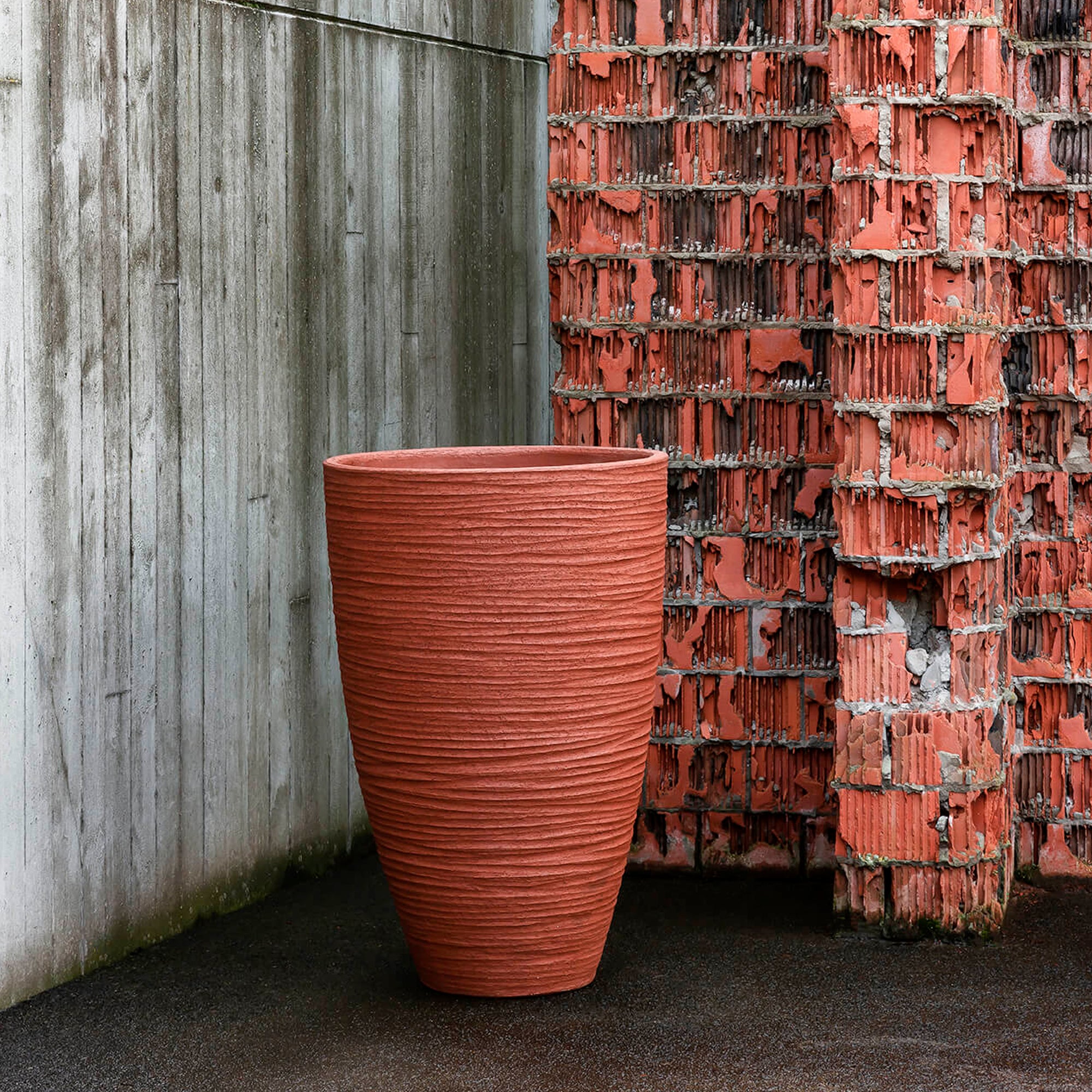 Eine rote Domani Vase Ø80cm mit einer Höhe von 135cm steht auf einer Holzterrasse vor einer Holzwand.