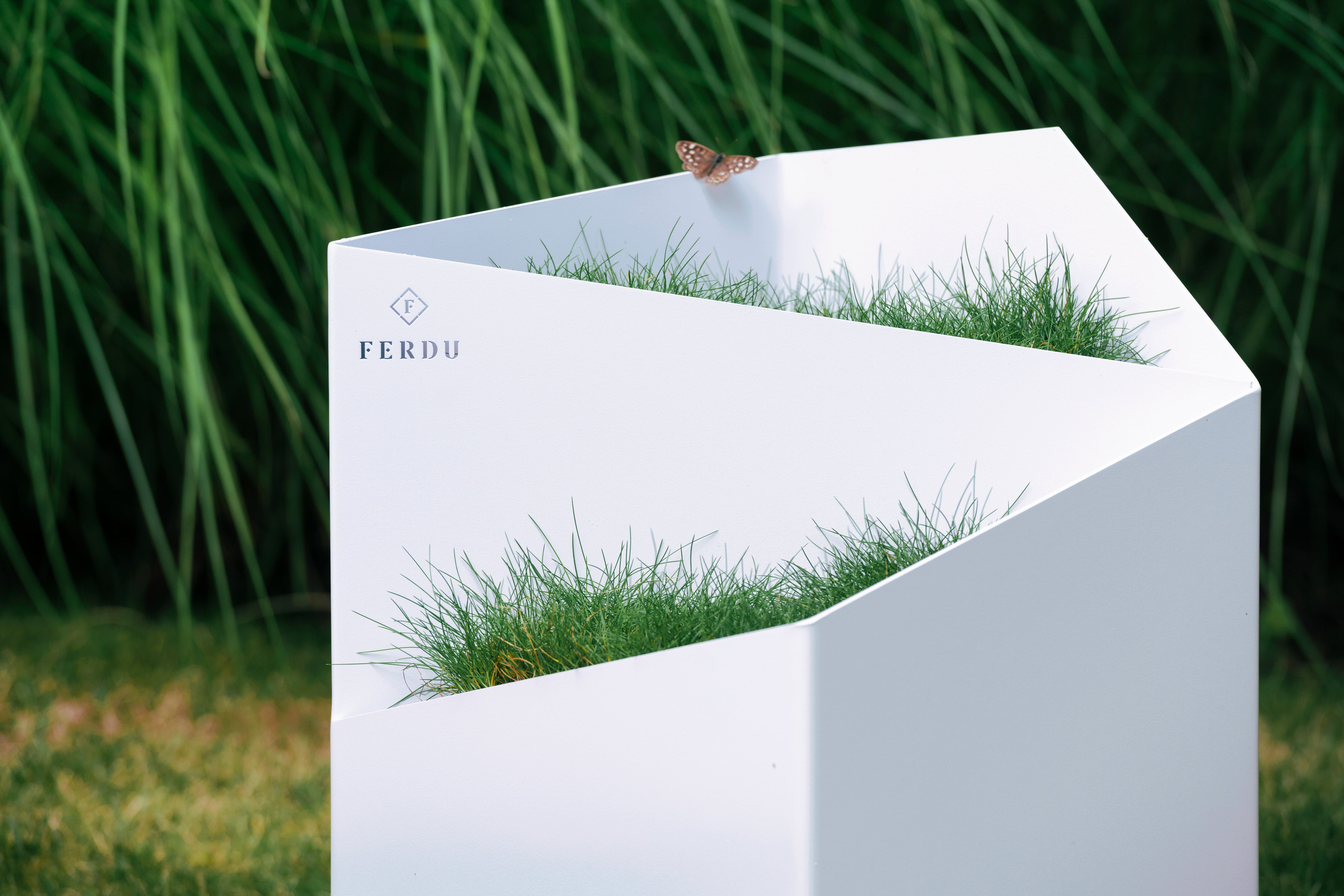 FERDU Gefaess Doblu aus CORTENSTAHL weiß lackiert auf einem Rasen. Im Hintergrund sind Gräser gepflanzt. Auf dem Rand sitzt ein  Schmetterling.