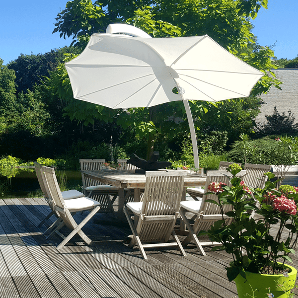 Umbrosa Sonnenschirm Icarus mit weißer Bespannung steht auf einer Terrasse. Unter diesem Design Schirm steht ein Esstisch mit mehreren Stühlen aus Teak.