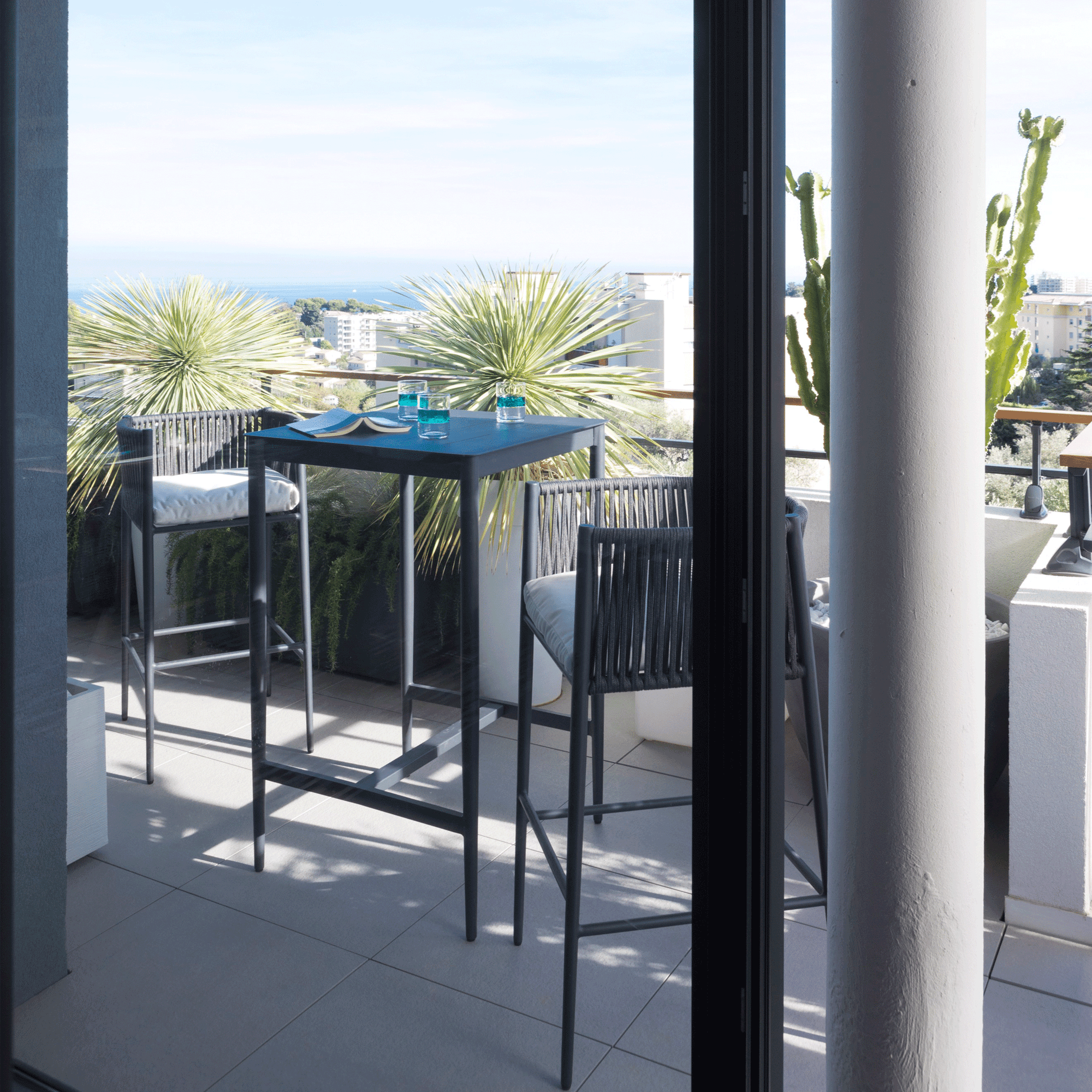 Auf einer Terrasse mit einem Blick auf Das Mittelmeer stehen zwei Barstühle und einquadratischer Bartisch der Kollektion Luce von Unopiu. Diese Gartenmöbel sind in grafitgrau und die Sessel sind mit weißen Sitzkissen ausgestattet.