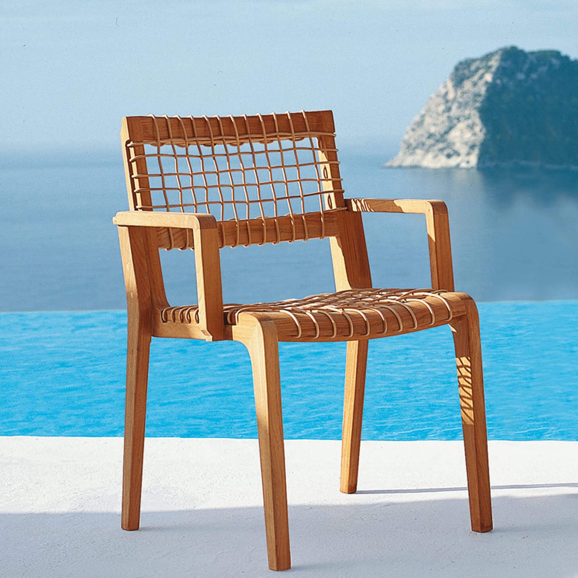 Unopiu Armstuhl Synthesis steht am Meer. Der Armstuhl ist aus Teakholz und hat die Maße 54x53x80cm.