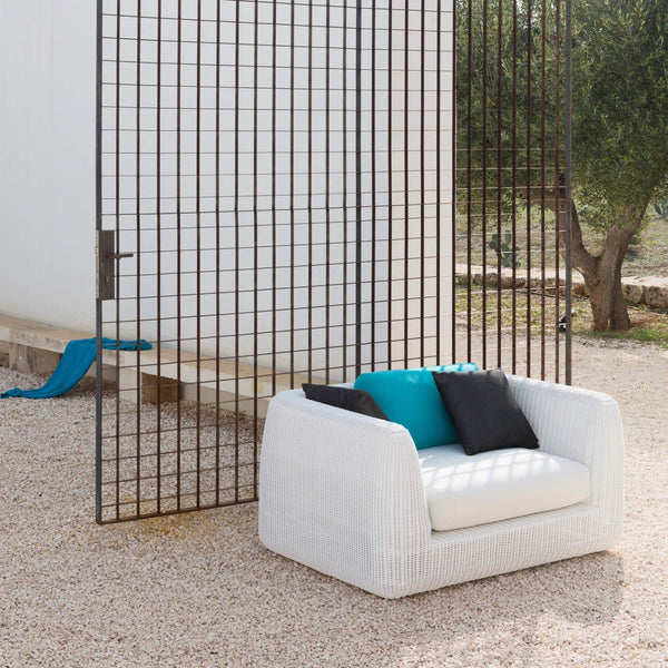 Unopiu´s weiß geflochtener Lounge Sessel steht in einem Garten  vor einem Olivenbaum. Als Rückenkissen befinden sich zwei braune Kissen und ein türkisfarbenes Kissen. Alle Kissen haben eine Größe von 40c40cm.