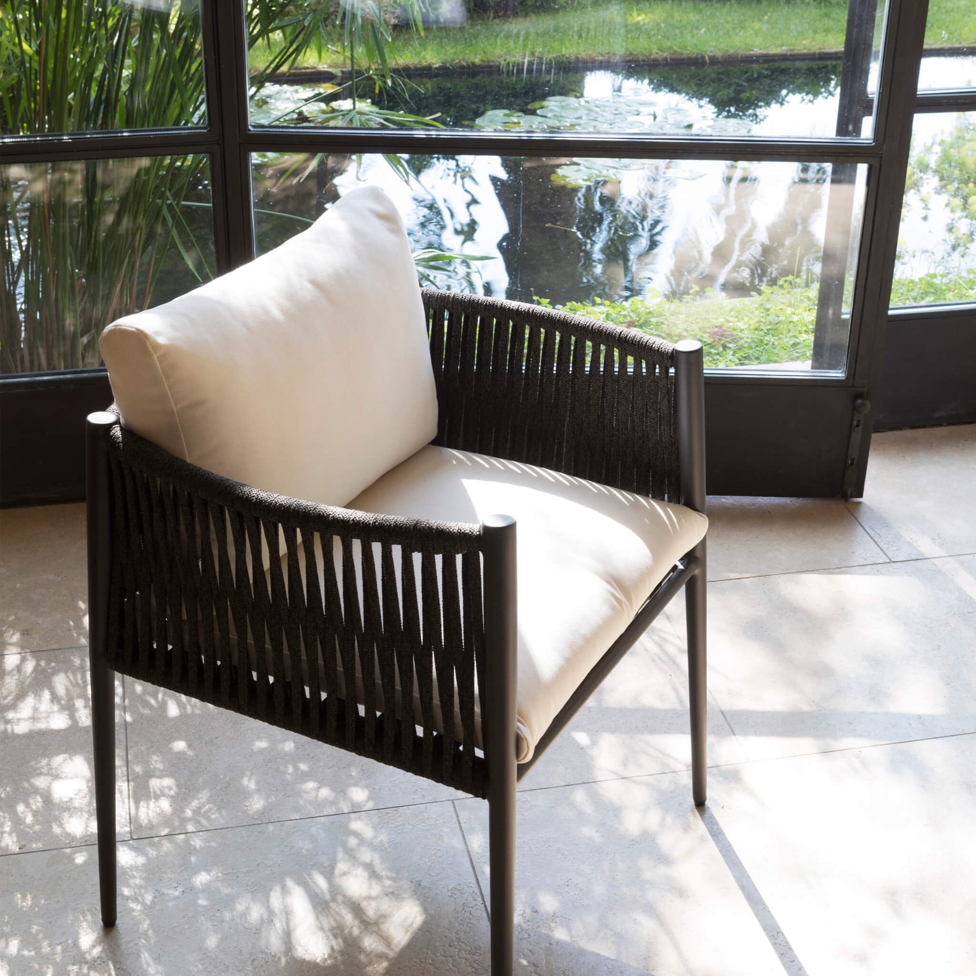 In einem Wintergarten steht der Unopiu Luce Sessel vor einer angewinkelten Glastür. Von diesem sehr bequemen Stuhl mit weisen Polstern schaut man auf einen kleinen Fluß der durch eine Wiese läuft.