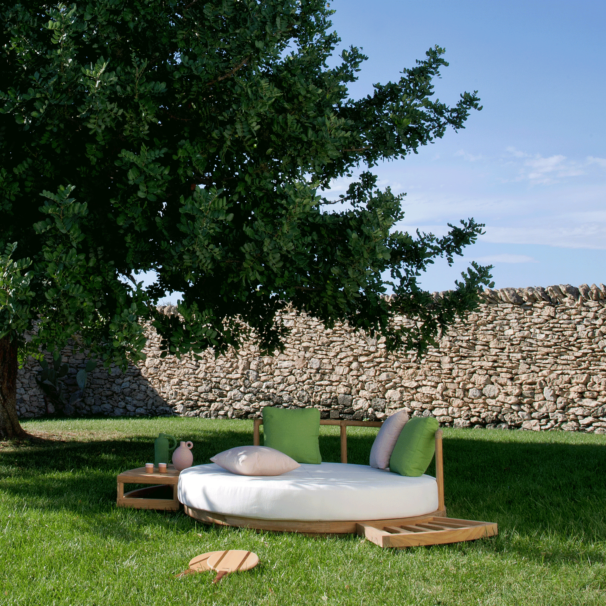 Unopiu Lounge Insel Pevero aus rundem Teakholz. Liegepolster in Acrylstoff weiß. Das Daybed steht im Rasen unter einem Haselnussbaum.